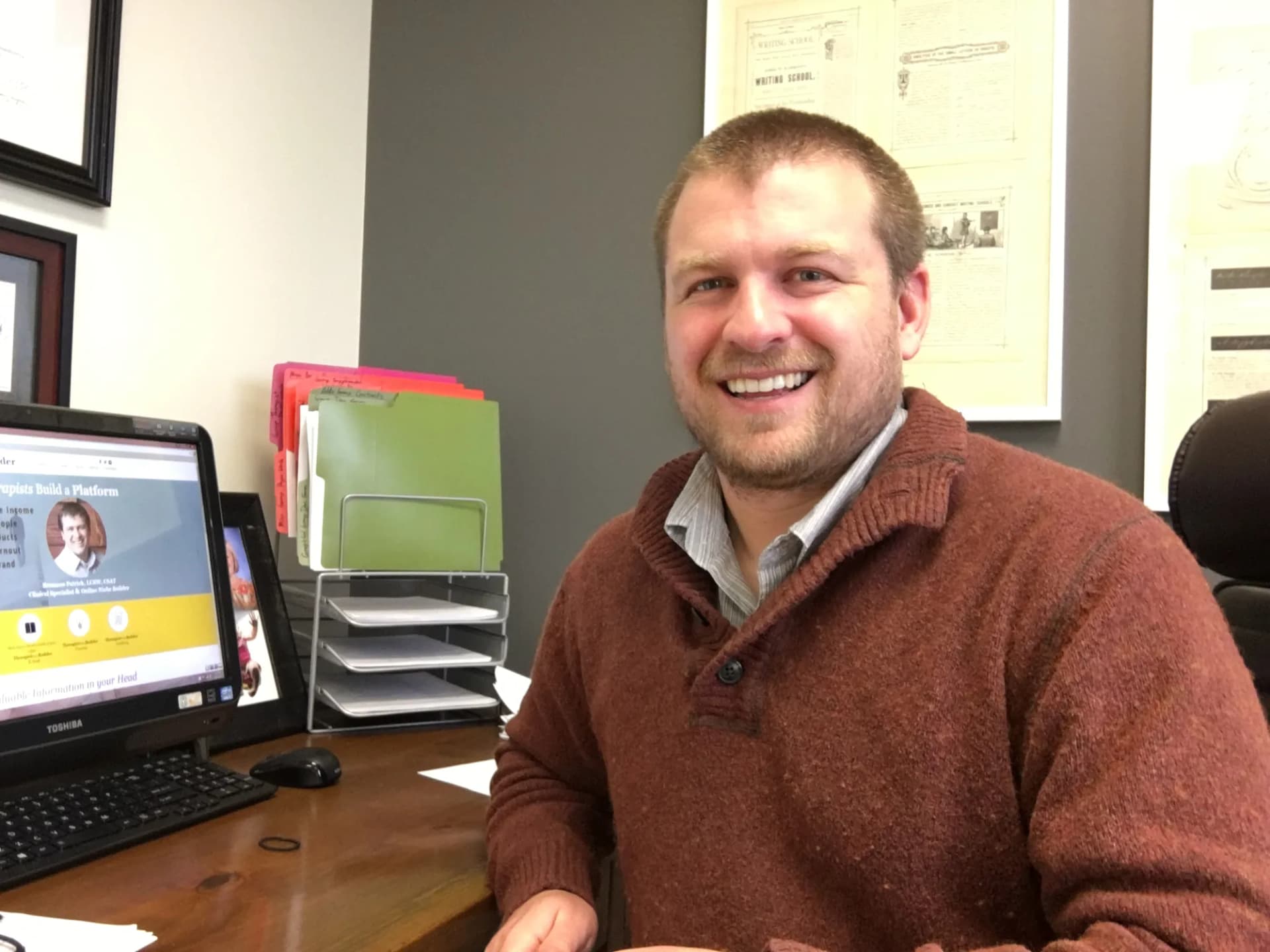 A younger Brannon at an office desk, smiling, with a presentation slide on the monitor beside him that reads Brannon Patrick, LCSW, CSAT.