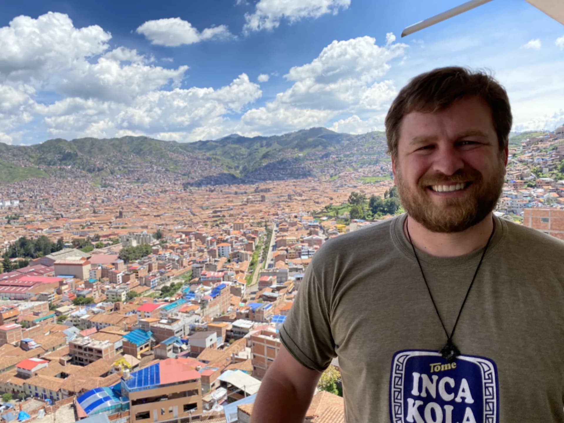 Brannon smiling on a hillside above a city of red tiled roofs, with green mountains and cloud behind him.