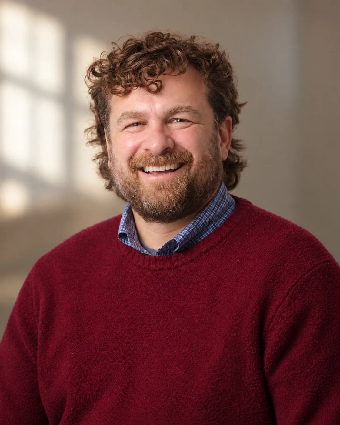 Brannon in a red sweater and checked shirt, smiling, in a room with light coming through a window behind him.