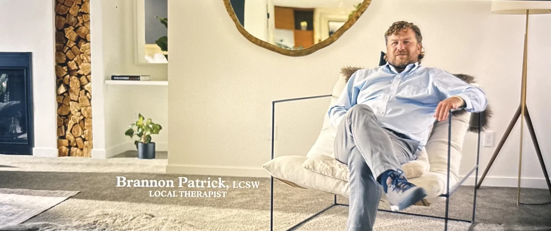 A still from the documentary: Brannon sitting in a low armchair in a bright room, with a caption on screen reading Brannon Patrick, LCSW, Local Therapist.