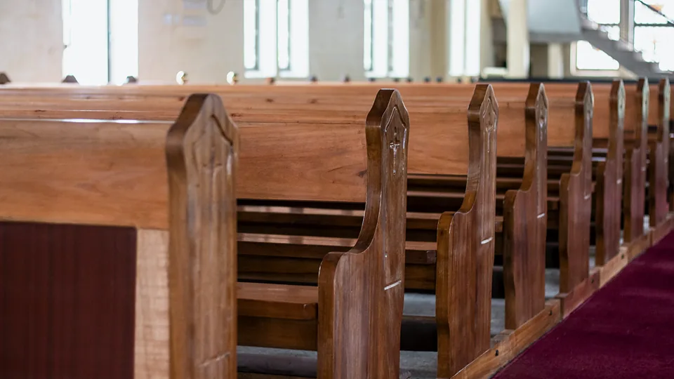 Rows of empty wooden pews in a chapel, lit from the windows behind them.