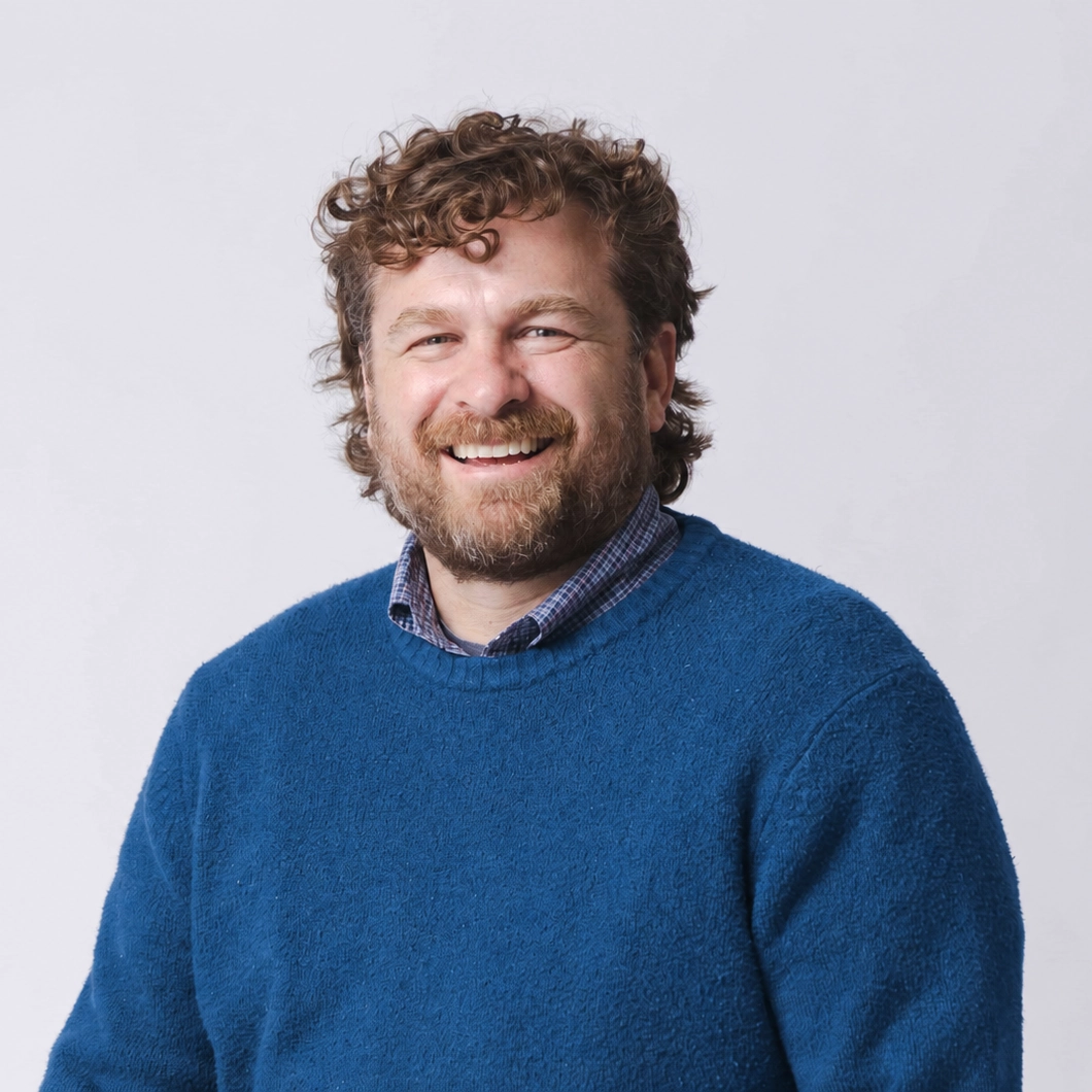 Brannon Patrick smiling, in a blue sweater over a checked shirt, against a plain light background