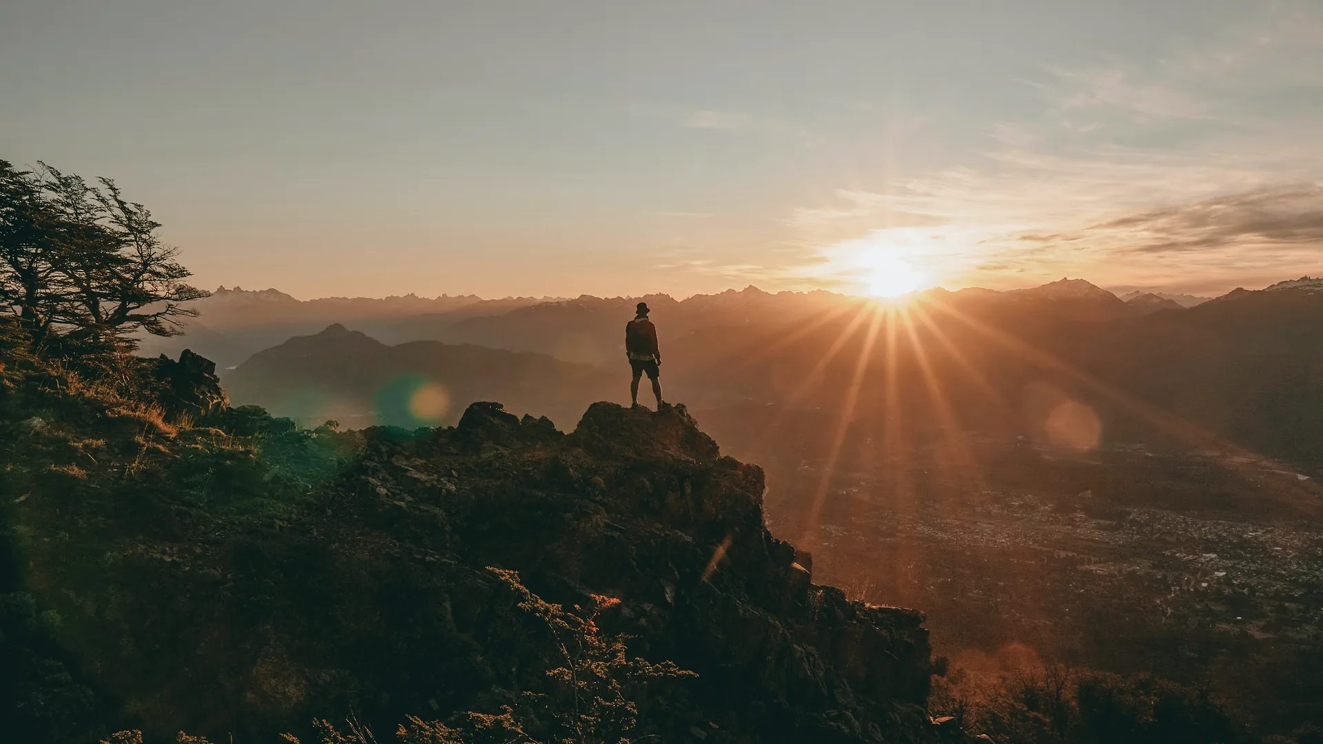 One person standing on a rocky summit, facing the sunrise over a valley.