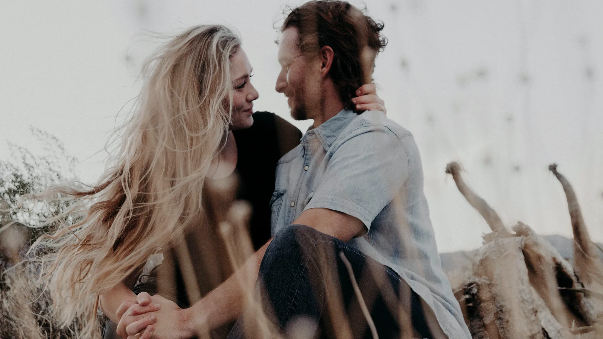 A couple sitting close together in tall dry grass, looking at each other.