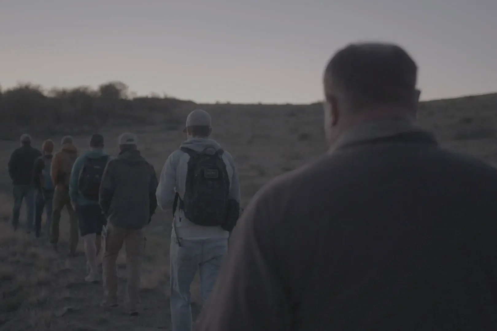 A line of men walking single file across open high desert at dusk, seen from behind.