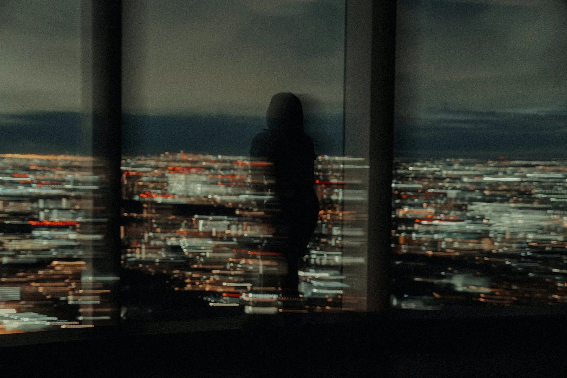 A person standing alone at a tall window above a city at night, seen in silhouette through the glass.