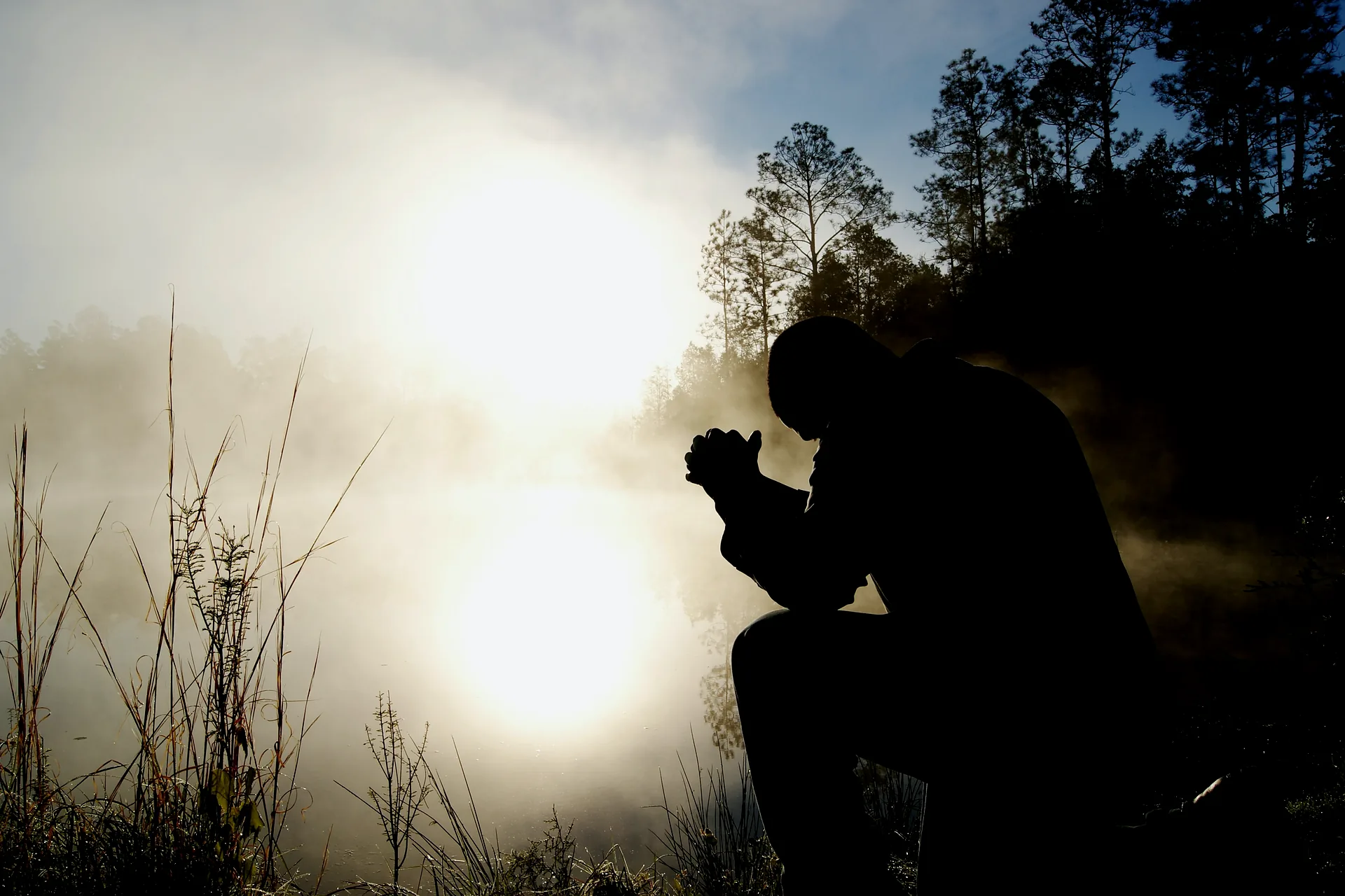 A man kneeling in silhouette beside a misty lake at sunrise, hands together and head bowed.