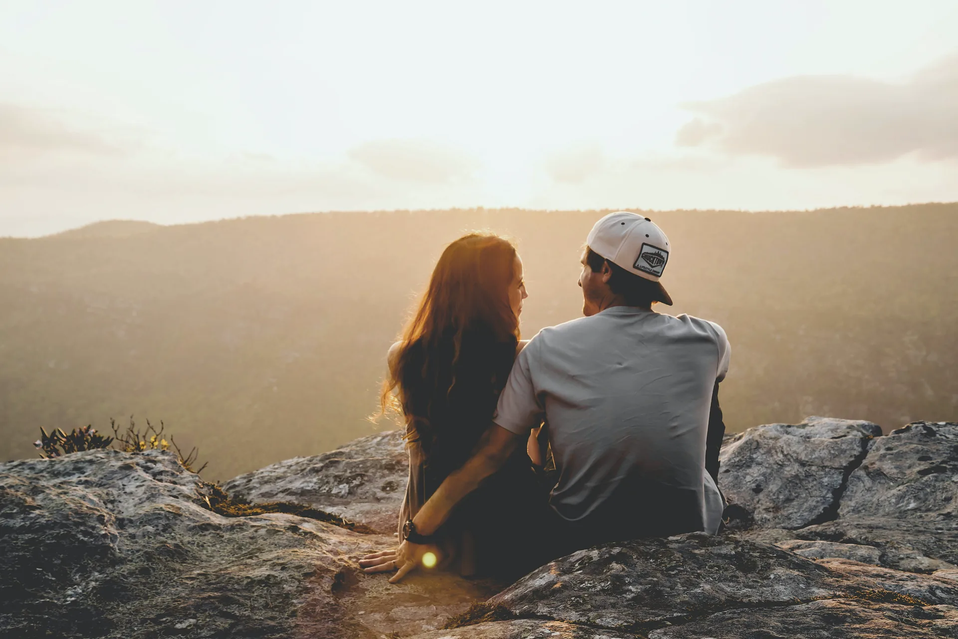 A man and a woman sitting close together on a rock at sunset, looking out over wooded hills.